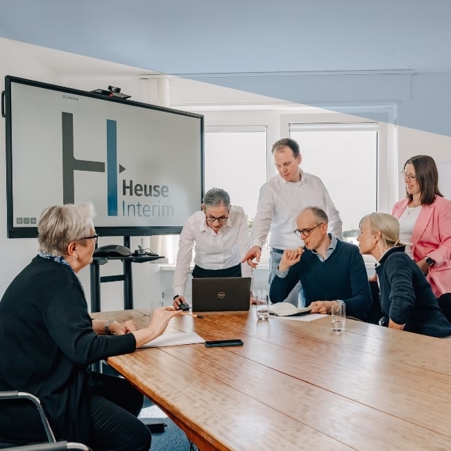 Team von Heuse Interim in einem Besprechungsraum, sechs Personen sitzen und stehen um einen großen Holztisch, eine Person zeigt auf einen Laptop, im Hintergrund ist ein Bildschirm mit dem Heuse Interim Logo zu sehen Team von Heuse Interim in einem Besprechungsraum, sechs Personen sitzen und stehen um einen großen Holztisch, eine Person zeigt auf einen Laptop, im Hintergrund ist ein Bildschirm mit dem Heuse Interim Logo zu sehen
