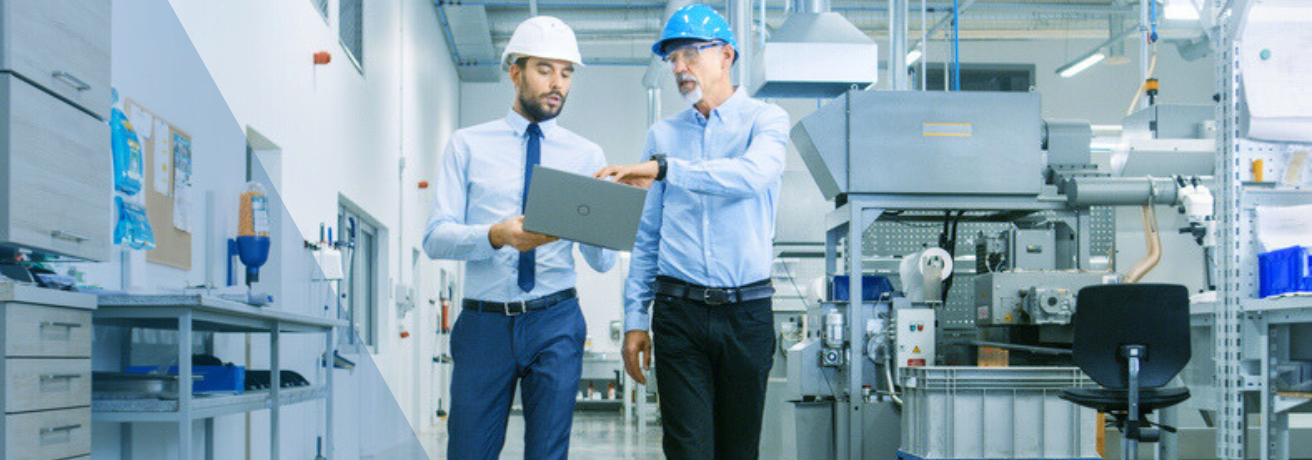 Zwei Männer mit Schutzhelmen gehen durch eine Produktionshalle und betrachten gemeinsam einen Laptop. Der ältere Mann zeigt auf den Bildschirm und erklärt etwas, während der jüngere aufmerksam zuhört; im Hintergrund sind Maschinen und technische Anlagen zu sehen.
