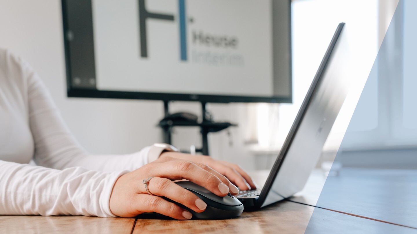 Hand einer Person an einer Computermaus neben einem Laptop, im Hintergrund ein Bildschirm mit dem Heuse Interim Logo im Büro von Heuse Interim
